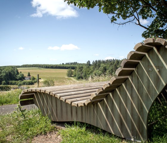 Holzliegebank mit Blick auf grüne Felder und Wälder unter blauem Himmel in Neidenbach., © TI Bitburger Land - Monika Mayer