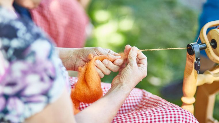 Een persoon spint oranje wol met een spinnewiel. Op de achtergrond zijn meer mensen te zien.