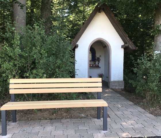 Eine kleine weiße Kapelle mit braunem Dach steht am Waldrand. Davor eine Holzbank auf gepflastertem Weg., © TI Bitburger Land