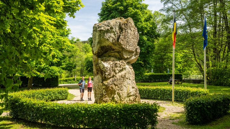 Two people are standing in front of a large rock in the green, flanked by two flags.