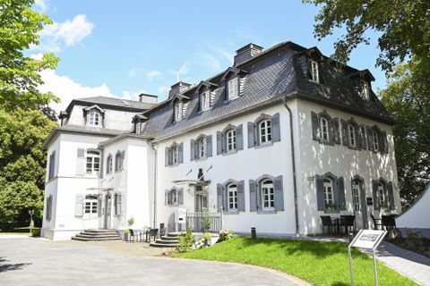 Ein elegantes Gebäude mit weißen Wänden und grauen Fensterrahmen steht inmitten eines gepflegten Gartens. Bäume und Sträucher umrahmen die Anlage.