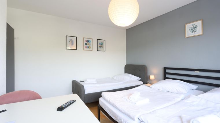 A simple room with two beds, a table, and a chair. Some pictures hang on the wall, and a lamp provides cozy light.