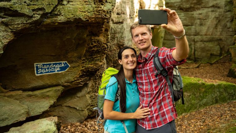 A couple takes a selfie in front of a rock entrance with a sign saying 'Mandrack Passage'.