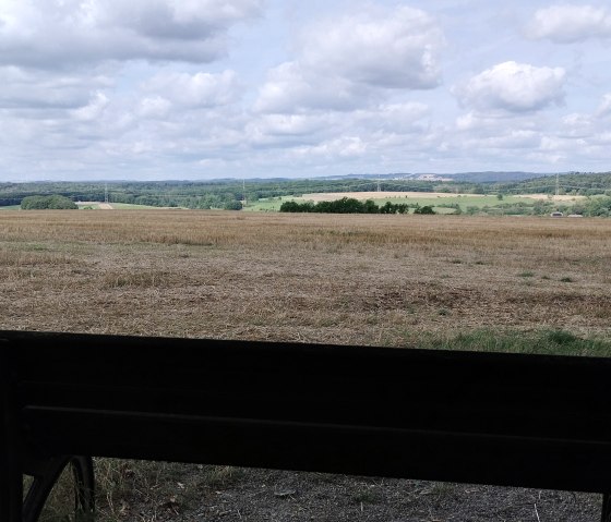 Eine Bank steht vor einem weiten Feld mit Blick auf bewaldete Hügel unter einem bewölkten Himmel., © TI Bitburger Land