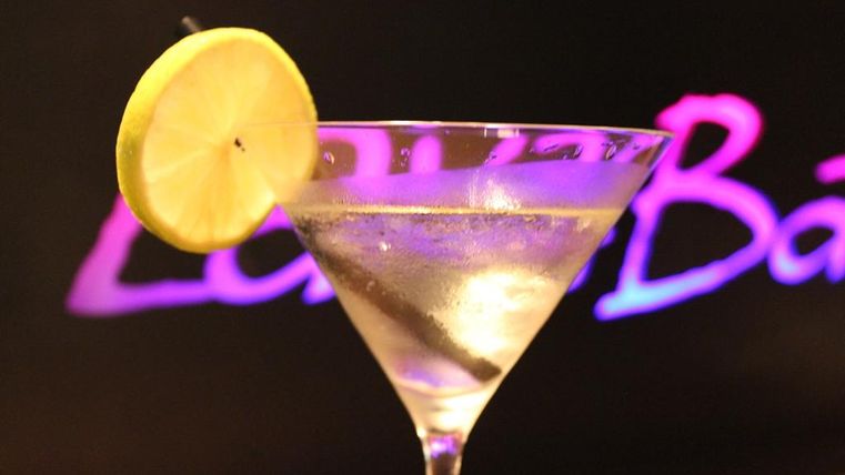 Een cocktail in een martini-glas met een schijfje citroen als garnering. Op de achtergrond straalt een kleurrijke tekst.