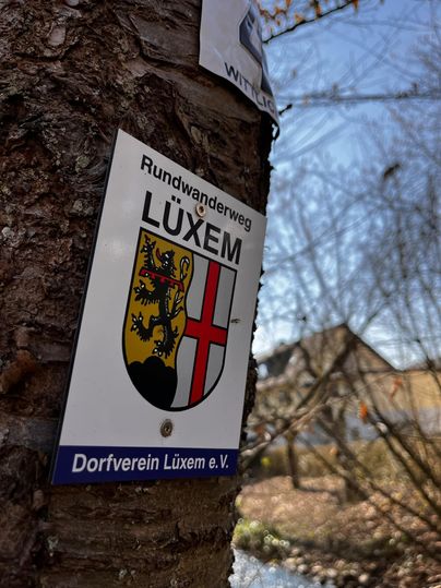 Een bord met het wapen van Lüxem toont de rondwandeling. Op de achtergrond zijn bomen en een blauwe lucht te zien.