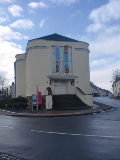 Ein rundes, modernes Gebäude mit einer hellen Fassade und großen Fenstern. Es steht an einer Straße mit einem klaren Himmel im Hintergrund.