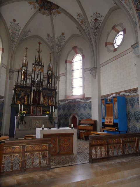 Ein Innenraum einer Kirche mit einem kunstvoll gestalteten Altar und bunten Fenstern. Auf der linken Seite steht eine Orgel und die Wände sind mit historischen Malereien verziert.