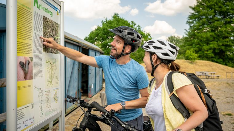 Two cyclists with helmets look at an information board on the Kyll cycle path.