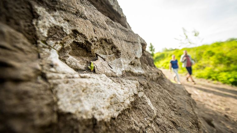 Close-up of a rock wall, where the volcanic layers can be seen. In the background, two hikers are walking towards the camera on the path.