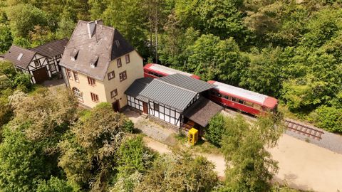 A red train is standing next to an old train station building amid a lot of greenery.
