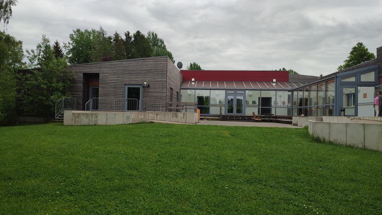 Un bâtiment moderne avec une façade en verre et une zone verte devant. Le ciel est nuageux et l'environnement semble calme.