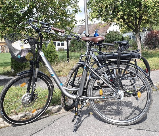 Deux vélos sont garés le long d'une route à Ingendorf. Un panneau indique la direction de Bettingen. En arrière-plan, on aperçoit des arbres et des maisons., © TI Bitburger Land, Steffi Wagner