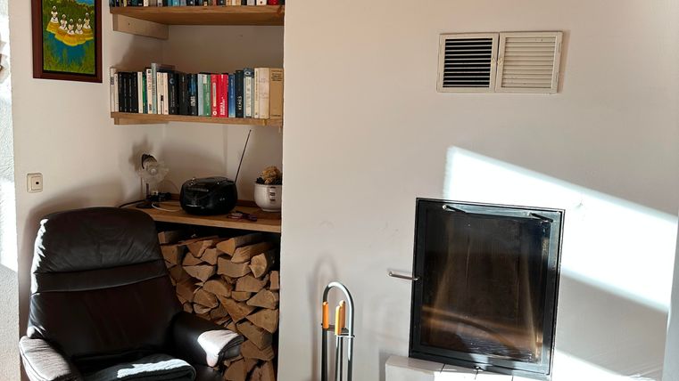 A cozy living space with a black armchair and a fireplace. In the background, a bookshelf and a woodpile are visible.