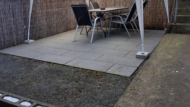 Een gezellige tuinruimte met een witte paviljoen en een tafel met stoelen. De vloer is betegeld en er is een muur van houten elementen.