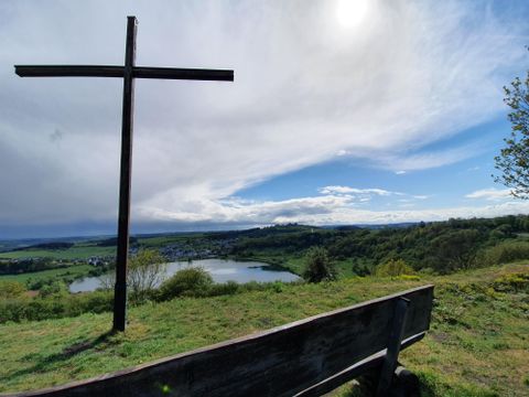 Un pont en bois mène à travers un lac tranquille, entouré d'herbe verte et d'arbres. Le ciel est clair avec quelques nuages.