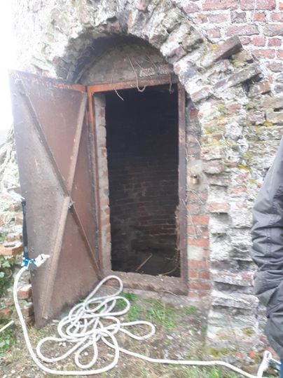 An old, open metal door leads into a dark room. A few hoses lie on the floor in front of the door.