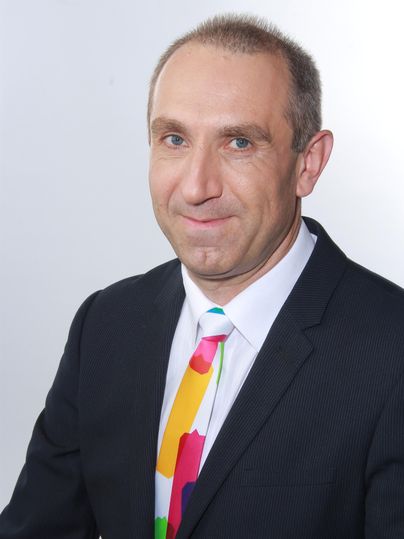 Ein Mann in einem Anzug mit buntem Krawatte lächelt in die Kamera. Der Hintergrund ist hell und neutral.