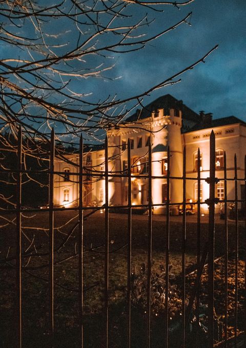 Een imposant kasteel dat 's nachts verlicht is. Op de voorgrond is een smeedijzeren hek en enkele takken zichtbaar.