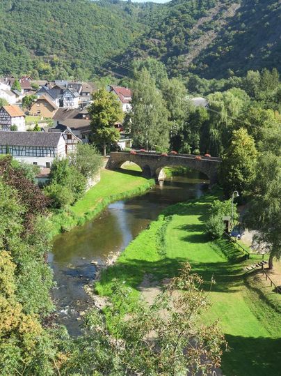 Un paysage pittoresque avec une rivière qui traverse un petit village. En arrière-plan, on peut voir des collines verdoyantes et des arbres.