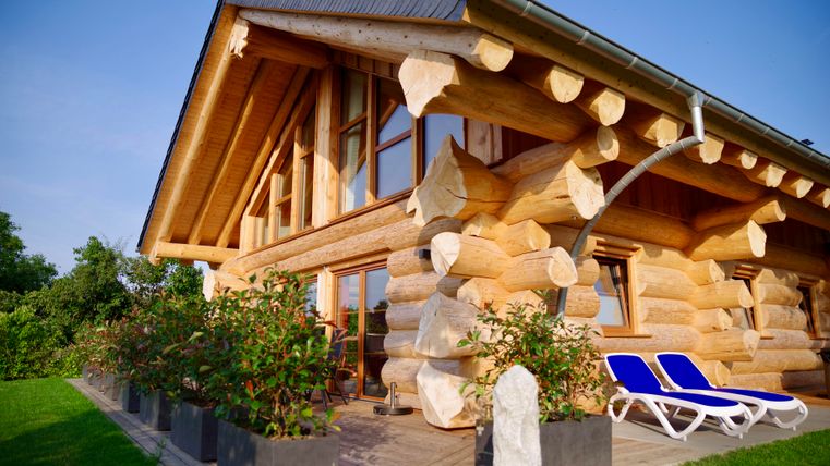 Une jolie maison en bois avec de grandes fenêtres et un toit moderne.
Au premier plan, il y a deux chaises longues et quelques plantes en pot.