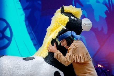 A man is hugging a large, plush horse with a yellow mane. The background is colorful and bright.