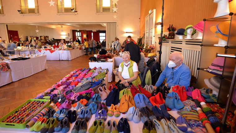 A marketplace filled with colorful shoes and accessories. Two elderly people are sitting in the foreground, while more vendor stalls can be seen in the background.