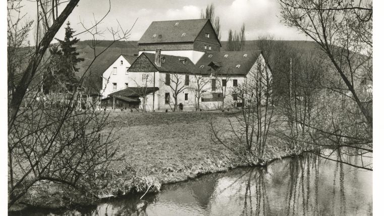 An old mill by the bank of a calm river. In the background, trees and a cloudy sky are visible.