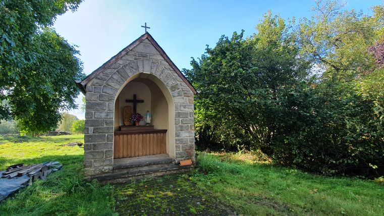 Kleine Steinkapelle mit Kreuz und Marienstatue, umgeben von Bäumen und Gras.