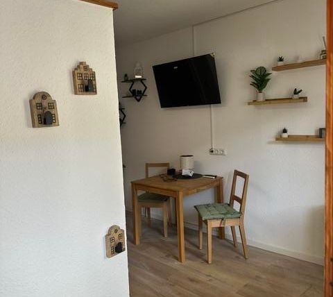 A cozy living space with a dining table and chairs. On the wall, there are shelves with plants and decorations.