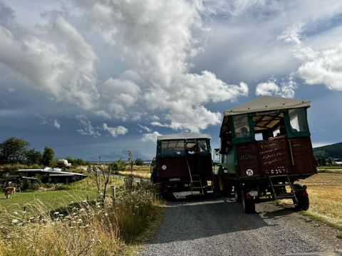 Two motorhomes are parked on a gravel road. In the background, fields and a cloudy sky can be seen.