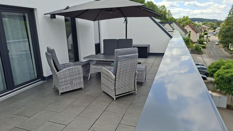 Eine moderne Terrasse mit einem Esstisch und Stühlen aus Rattan. Ein großer Sonnenschirm spendet Schatten und im Hintergrund sind einige Häuser zu sehen.