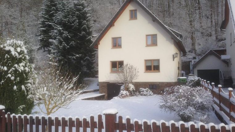 A pretty house in the snow, surrounded by a fenced garden. In the background, snow-covered trees can be seen.
