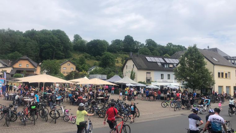 A lively scene with many people and bicycles in a square. In the background, houses and trees can be seen.