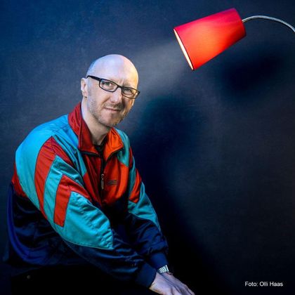 Ein Mann mit Brille sitzt neben einer roten Lampe. Er trägt eine bunte Trainingsjacke und lächelt in die Kamera.