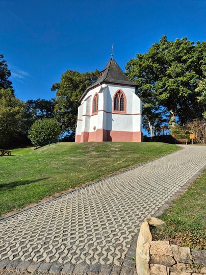 Eine kleine Kapelle steht auf einem Hügel, umgeben von Bäumen und grünem Gras. Der blaue Himmel verleiht der Szene eine ruhige Atmosphäre.
