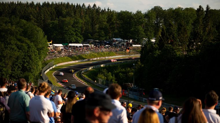 A large crowd is watching a racetrack as cars speed around the corners. Trees and spectator stands can be seen in the background.