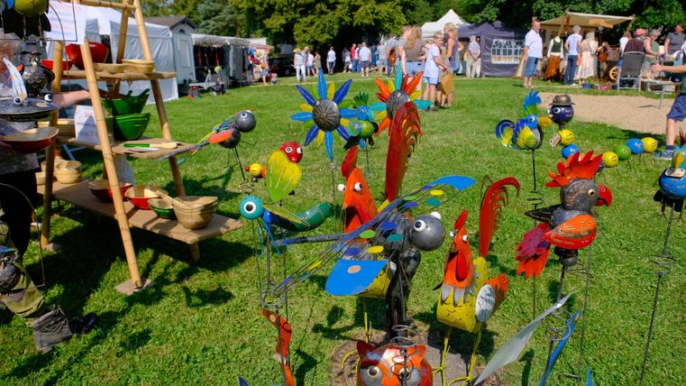 Bunte Gartenfiguren aus Metall stehen auf einer Wiese. Im Hintergrund sind Menschen auf einem Markt zu sehen.