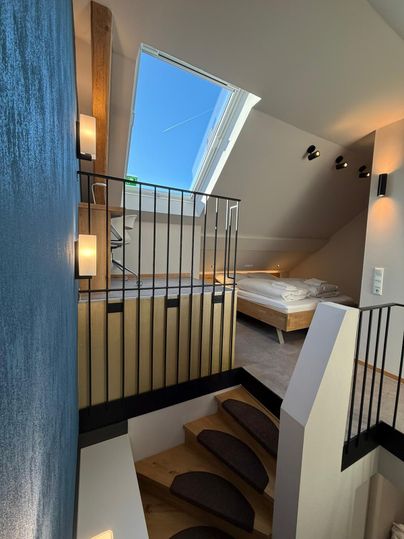 A modern interior with a wooden staircase and bright light. Upstairs is a cozy bedroom with a large window.