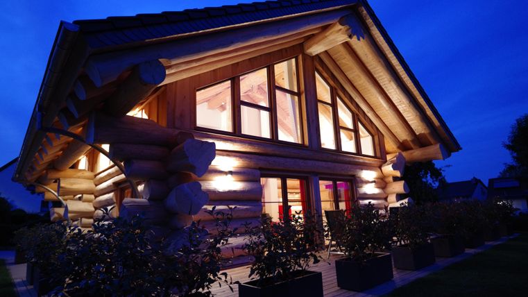 Une maison en bois moderne avec de grandes fenêtres et une lumière chaude. Elle a une atmosphère accueillante la nuit.