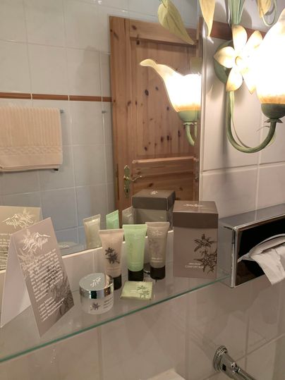 Een badkamer met verschillende verzorgingsproducten op de wastafel. Op de achtergrond zijn een houten deur en decoratieve lampen zichtbaar.