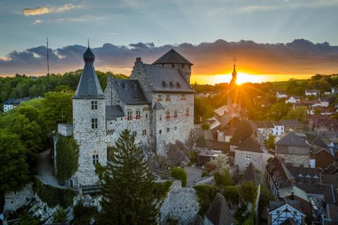 Eine beeindruckende Burg umgeben von Bäumen und Häusern. Im Hintergrund geht die Sonne auf und taucht die Landschaft in warmes Licht.