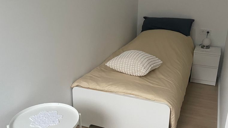 Ein schlichtes Schlafzimmer mit einem Einzelbett und einer weißen Nachttischlampe. Der Raum hat einen hellen Boden und neutrale Wände.