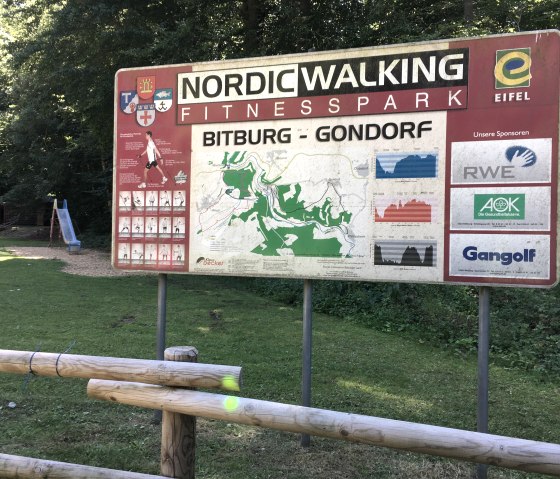Schild des Nordic Walking Fitnessparks Bitburg-Gondorf mit Karte und Sponsorenlogos im Wald, daneben ein Spielplatz mit Rutsche., © TI Bitburger Land