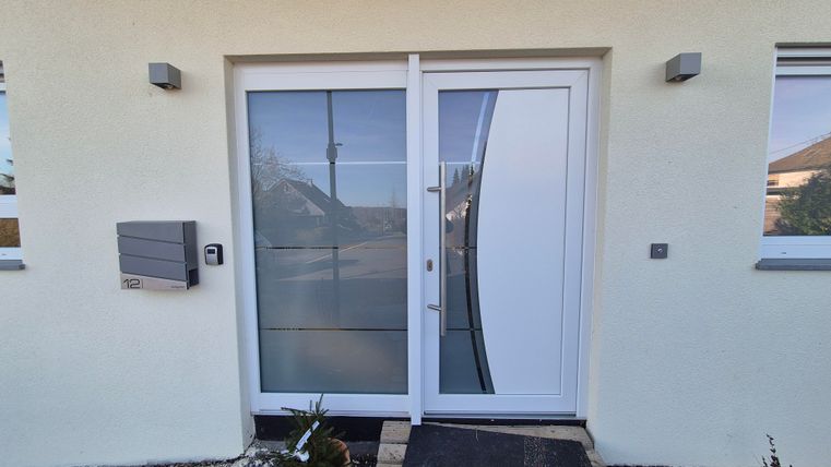 Une entrée moderne avec une porte en verre élégante et un paillasson simple. À gauche de la porte se trouve une boîte aux lettres et quelques plantes vertes devant le bâtiment.