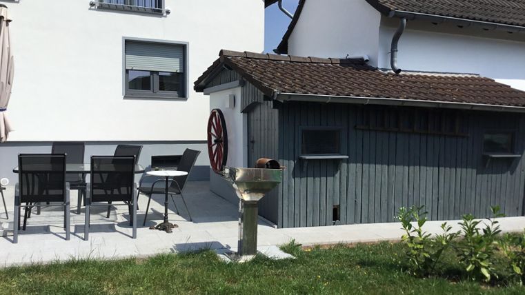 A beautiful garden with a small terrace and modern garden furniture. In the background, there is a wooden shed with a rustic wheel.