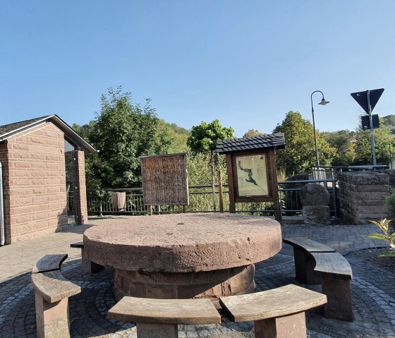 Place de village à Brecht avec un banc rond, des panneaux d'information et un petit bâtiment. Entourée d'arbres et d'une route en arrière-plan., © TI Bitburger Land, Steffi Wagner