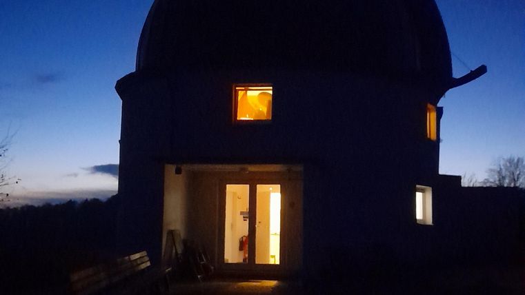 Een observatoriumachtig gebouw 's nachts, verlicht van binnen. De lucht is donkerblauw en helder.