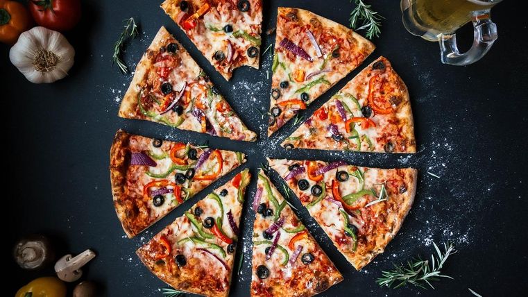 Pizza in acht stukken met groenten en olijven, omringd door tomaten, knoflook en een bierglas op een zwarte achtergrond.