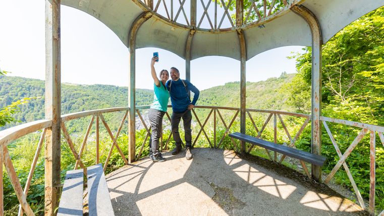 Zwei Personen machen ein Selfie in einem Pavillon mit Blick auf bewaldete Hügel.